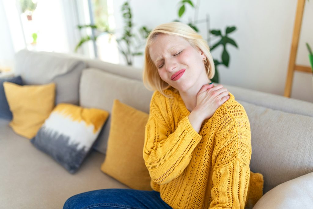 A woman in a yellow sweater sits on a couch, massaging her neck and shoulder, appearing to manage chronic pain effectively.