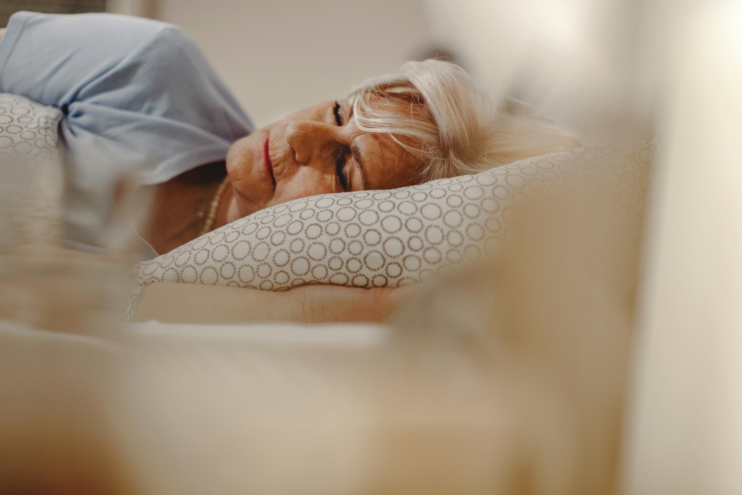 An elderly person with gray hair, possibly free from sleep disorders common in their age group, is sleeping peacefully on a pillow with a geometric pattern. They're wearing a blue shirt, and the scene is softly lit.