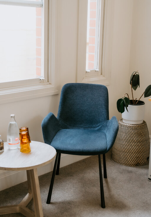 A blue chair is next to a small round table with two amber glasses and a bottle. A plant is in a woven basket nearby, in a room with beige walls and windows.